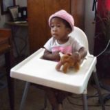 Photo of a toddler girl sitting in a high chair. She has brown skin; she is wearing a white shirt and pink cap, and holding a teddy bear in one hand.