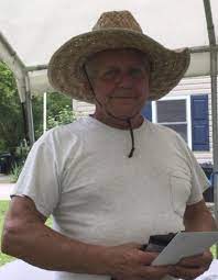 Photo of an older man with gray hair and tanned skin, wearing a white T-shirt that stretches over a beer belly and a straw hat with a leather tie around the chin.
