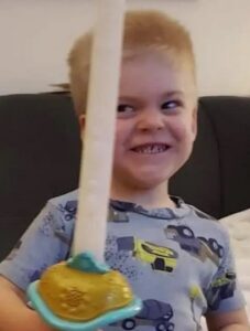 Photo of light-skinned, blond boy wearing a blue print shirt and grinning. He is holding a plastic toy sword.