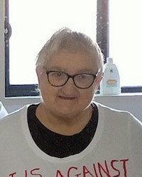 Photo of an older woman with short white hair, pale skin, and a round face. She is wearing black plastic-rimmed glasses and a white sweater over a black shirt.