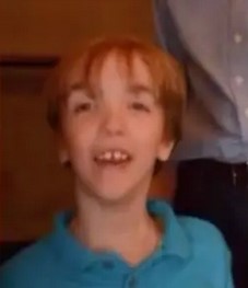 Blurry photo of a young boy with pale skin, red hair cut into bangs that hang over his eyes. His chin is pointed, and he is smiling, showing a gap between his front teeth. He is wearing a teal polo shirt.