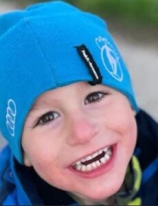 Photo of a young boy with pale skin and dark-brown eyes, wearing a bright blue cap and yellow-and-blue jacket, smiling at the camera.