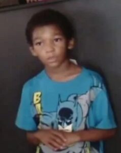 Photo of a solemn African-American boy with light-brown skin and short curly dark-brown hair, wearing a blue T-shirt that is a bit too big for him.