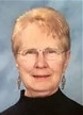 Photo of an elderly woman with fair skin and short blonde hair, wearing glasses, dangling earrings, and a black turtleneck. She is smiling for the camera.