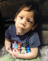 Photo of a boy sitting on carpet. He has light-brown skin and short straight black hair. He is wearing a black T-shirt with a colorful logo on the front. His expression is somewhat worried, and his head is tilted to the side.