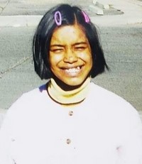 Photo of a girl with tan skin and straight, chin-length black hair, smiling fo the camera and squinting into the sun. She is wearing a white turtleneck sweater, and has pink barrettes in her hair.
