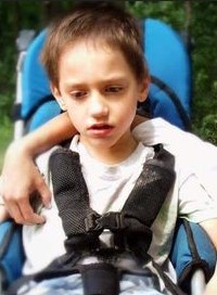 Photo of a boy with pale skin and short brown hair, wearing a white T-shirt and strapped into his chair with a four-point harness. His expression is serious.