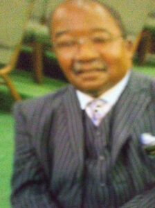 Photo of an older African American man wearing a gray tuxedo and glasses. He is smiling for the camera. His hair is a short fringe on the sides of his head.