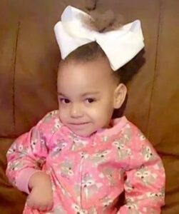 Photo of a toddler girl wearing a pink print long-sleeved shirt. Her skin is light tan and her hair, pulled into a huge white satin bow, is curly and brown; she is half-smiling for the camera, and one of her small hands is tucked in close to her body.