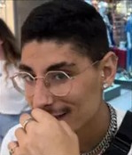 Photo of a young man with short black hair, small mustache, and light-brown skin. He is wearing wire-rimmed glasses and a black and white shirt. He has his hand to his mouth; there is a silver ring on his second finger. He is smiling slightly. 