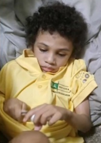 Photo of a boy with light-brown skin and curly brown hair, lying on his back. He is wearing a yellow shirt. His expression is pensive, and he is looking at something he is holding in his hands.