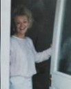 Photo of a woman with curly blond hair and fair skin, smiling and holding open a door; she is wearing a white dress.