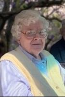 Photo of an older lady with pale skin and curly, short white hair; she is wearing square-rimmed glasses, a pastel purple striped blouse, and a yellow vest.