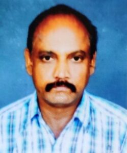 Portrait photo of a man with tan skin, black hair and bushy mustache, wearing a checked blue and white shirt.