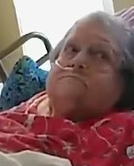 Photo of an elderly woman in a red shirt, lying in a hospital bed; she has fair skin and short gray hair. She is wearing an NG tube and looking off to the side.
