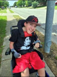 Photo of a teenage boy sitting in a wheelchair. He has pale skin; he is smiling. He is wearing a red and black baseball cap, a black T-shirt, red shorts, and gray fingerless gloves. He is smiling, head tilted forward and to one side.