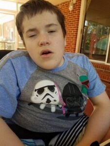 Photo of a teenage boy sitting in a wheelchair, head tilted to one side. He has pale skin and short brown hair, and is wearing a blue and gray shirt with the image of a Star Wars Storm Trooper and Darth Vader on it. His eyes are half-closed, and he looks sleepy.