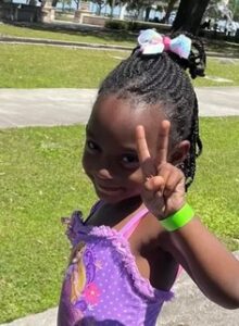 Photo of a small girl with her hair in braids and a bow atop her head, smiling for the camera and holding up a "V" sign. She is wearing a purple tank top and a lime-green bracelet.
