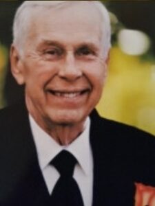 Photo of a smiling older man in a black suit and tie; he has light skin and short white hair.