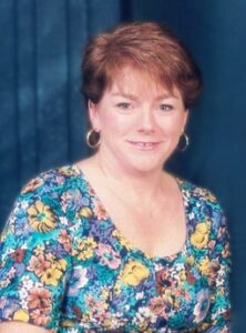 Photo of a woman with short auburn hair and pale, sunburnt skin, wearing a flowered dress.