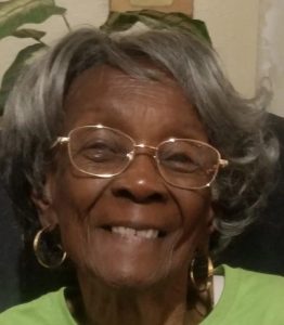 Photo of an older woman with deep brown skin and wavy gray hair; she is smiling for the camera. She is wearing gold-rimmed glasses and gold hoop earrings.