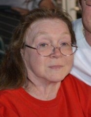 Photo of an older woman with pale skin and brown hair pulled back from her face; she is wearing glasses, which are slipping down her nose, and a bright red sweater.