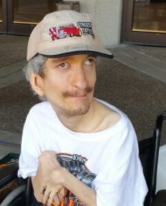 Photo of a man sitting in a wheelchair. He is wearing a white T-shirt printed with a logao and a worn beige baseball cap. He has light skin, fuzzy gray hair, and a sparse mustache and beard. One of his eyes is pointed off to the side, and his hands are thin and bent with contractures.