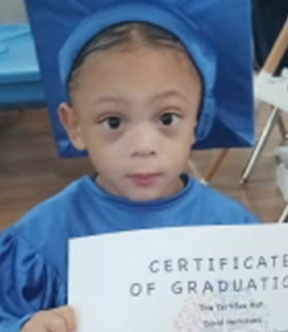 Photo of a child in a blue graduation gown and mortarboard, holding a diploma. He has dark hair and light-brown skin.