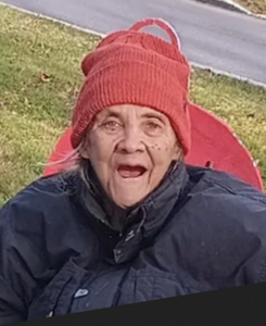 Photo of an elderly woman with light skin, wearing a bulky brown jacket. Wisps of hair poke out from under a red knit cap.
