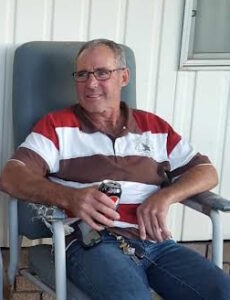 A fair-skinned man in late middle age sits in a gray-upholstered lawn chair, holding a can of soda. He is wearing glasses and a red-and-white striped polo shirt. 