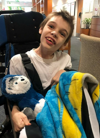 Photo: A smiling boy sitting in a wheelchair, holding a stuffed teddy bear. He has pale skin and short brown hair. A yellow and blue blanket is draped across his lap.