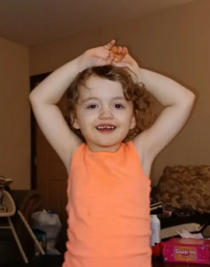 Photo of a small girl with pale skin and short, messy red hair, smiling. She has her hands clasped above her head and is wearing an orange tank top.