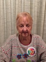 Photo of an older woman with gray hair; she has pale, wrinkled skin and is wearing a beige flowered sweater and a pin with the number ninety on it.