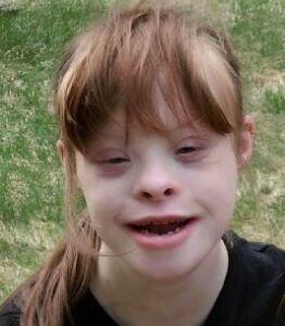 Photo of a girl with pale skin and straight auburn hair. She has Down syndrome. She is smiling broadly.