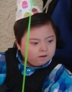 Photo of a round-faced boy with Down syndrome, wearing a blue shirt and cupcake party hat. He has soft brown hair and fair skin. 