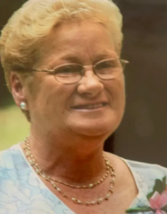 Photo. An older woman with short, permed blond hair and fair skin, wearing glasses, pearl earrings and necklace, and a blue top. She is smiling for the camera.