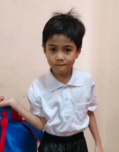 Photo of a small boy wearing a collared shirt and black pants, his black hair cut short and sticking up a little, holding a blue and red backpack. He has tan skin and deep brown eyes.