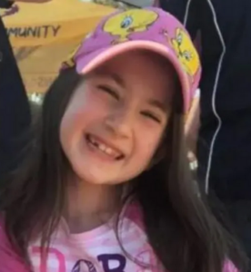Photo of a young girl with light skin and dark-brown hair tucked under a pink Tweety Bird ball cap. She is grinning, head tilted to one side. She is wearing a pink T-shirt.