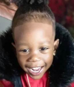 Photo of a small boy with light-brown skin and brown eyes. He is grinning. He is wearing a red coat with a fuzzy black hood.