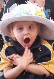 Photo of a small girl with fair skin and blond hair, wearing a large white flowered hat and an oversized blue and yellow sweatshirt. She has her mouth open and is reaching for the camera.