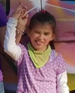 A young girl with light skin and fine brown hair tied back in a pink hair tie; she is wearing a pink top and yellow bandana. She is winking and sticking her tongue out at the camera. She is holding up her hand, which is being held by someone off-frame.