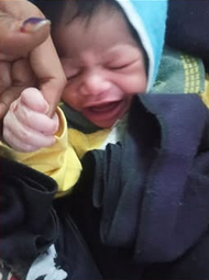 Photo of a newborn baby, face scrunched up. She is grabbing the hand of an adult. She is wrapped in blankets. Her skin is light-brown.