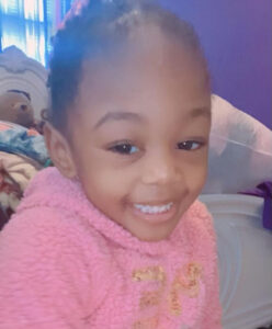 Photo of a smiling toddler wearing a pink sweater. She has light-brown skin and her dark hair is pulled back into braids.