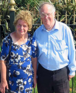 Photo of an elderly couple standing outdoors. On the left is a woman in a blue dress, with pale, somewhat sunburnt skin and curly blond hair; on the left is a balding man with a fringe of white hair, light skin, and glasses, wearing a blue dress shirt and black pants.