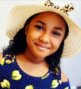 Photo of a young girl wearing a straw hat and blue dress. Her skin is light-brown; she has shoulder-length curly brown hair. She is smiling for the camera.