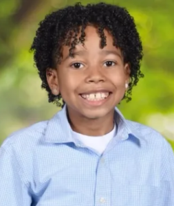 Photo of an African American boy with light-brown skin, his dark-brown hair in tight curls. He is wearing a blue dress shirt and smiling for the camera.