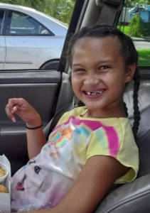 Photo of a young girl sitting in a car. She has light-brown skin, and her brown hair is done in two braids. She is wearing a tie-dyed T-shirt. She is smiling broadly, showing gaps between her teeth, and her eyes are squinted almost shut.