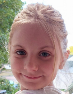 Photo of a girl with wispy blond hair tied back from her face; her skin is pink and she has large blue eyes. She is smiling.