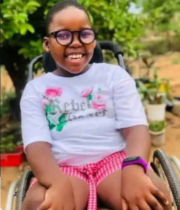 Photo of a grinning child sitting in a wheelchair. Her skin is brown; her black hair is pulled neatly back from her face; she is wearing a white shirt, red checked shorts, and a pink watch. 