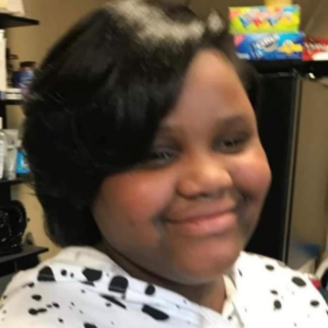 Photo of a teenage girl with a round face and straight black hair cut to chin length. She has brown skin, is wearing a white shirt with black spots, and is smiling for the camera.